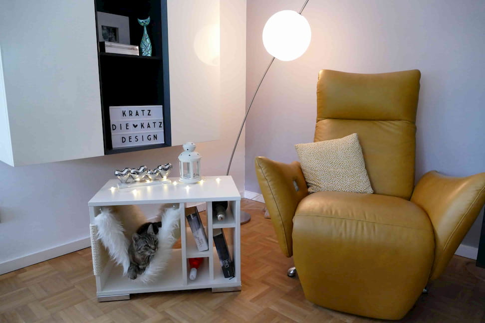 Katze liegt in der Hängematte eines breiten, weißen Kratzwürfels mit mehreren Regalfächern, in denen Bücher und Weinflaschen liegen und auf dem Blumen stehen. Der Kratzwürfel steht neben einem hellbraunen Sessel in einem modern eingerichteten Wohnzimmer.